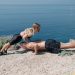 A man and woman perform outdoor fitness exercises on a cliff with a breathtaking sea view.